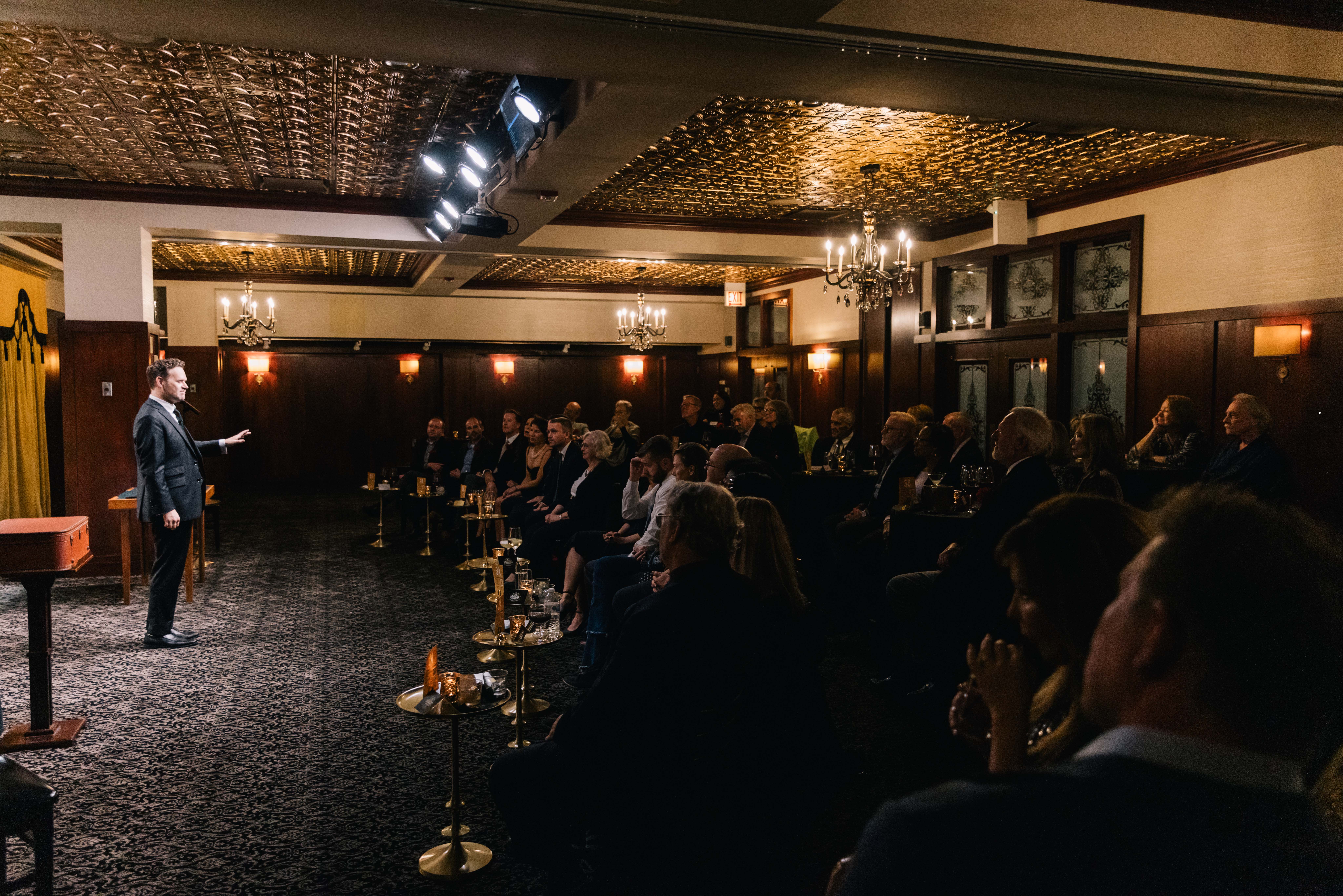 Dennis Watkins performing to a full audience in a room with low top and high top tables and chairs, gold curtains, and dark wood walls.